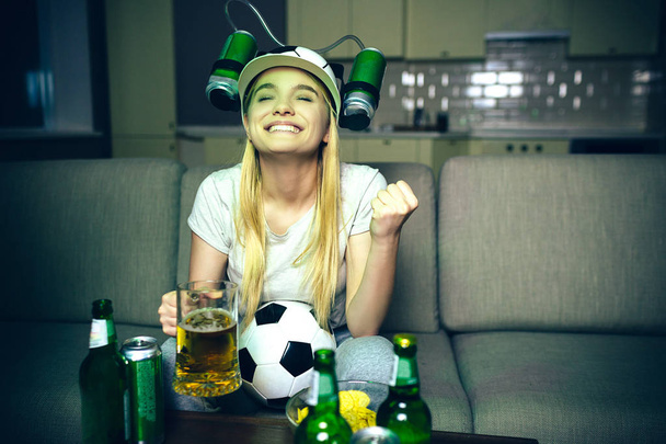 Young woman watch football game on tv at night. Cheerful positive model smiling. Holding hand in fist. Looking up. Glass of beer and bottles on table. Ball between legs. - Foto, imagen