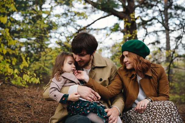 Young father, mother and little toddler daughter girl in a beret and a coat sitting on the ground in the autumn forest. - Foto, afbeelding