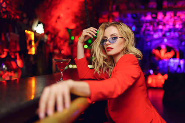 Fotografie mladé blonďaté ženy s brýlemi a červeným oblečením, sedící v baru s koktejlovou sklenicí v nočním klubu - Fotografie, Obrázek