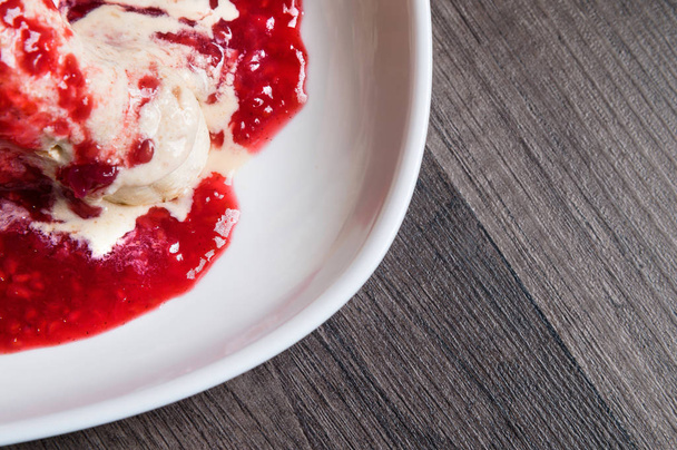 Closeup Vanilla ice cream poured with strawberry or raspberry jam with sliced banana slices on a white plate on a dark wooden table. You can do this at home on vacation - Фото, изображение