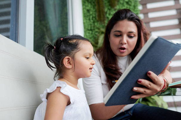 Giovane madre che tiene il libro e insegna alla sua piccola figlia a leggere mentre si siedono sul divano all'aperto
 - Foto, immagini