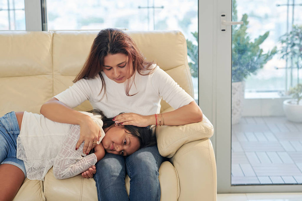 Linda chica acostada en las rodillas de su madre y su madre abrazándola mientras se sientan en el sofá en casa
 - Foto, imagen