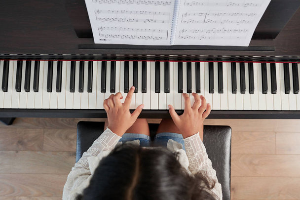 Вид зверху на молоду дівчину, яка сидить на фортепіано і грає музичну композицію під час уроку
 - Фото, зображення
