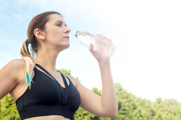 Portrait of young beautiful dark-haired fitness woman drinking water at summer green park. - 写真・画像