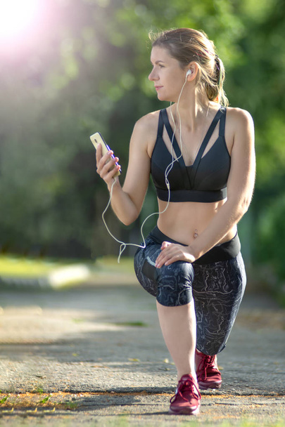 Front view woman is stretching in the city park and listens music on headphones. Fitness and lifestyle concept. - Zdjęcie, obraz