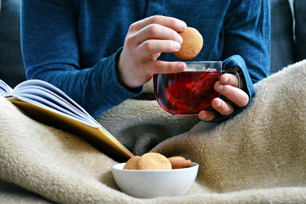 Un joven está sentado en un sofá y leyendo un libro mientras sostiene un té de frutas en una taza de vidrio en la mano y galletas a su lado - primer plano del libro y taza de vidrio llena de té - concepto para calentar
 - Foto, Imagen