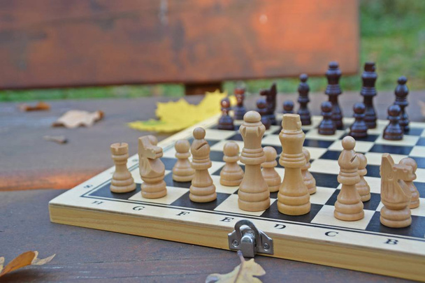 A sakktábla a sakk darab áll a fából készült asztal az őszi erdő színes őszi levelek mellette-két sakkfigurák szembe egymással, ami a harcot a megfelelő csapatok - Fotó, kép