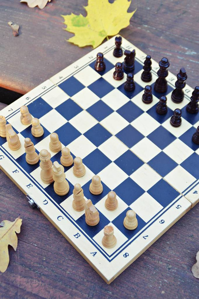 Ein Schachbrett mit Schachfiguren steht auf einem Holztisch im Herbstwald, daneben bunte Herbstblätter - zwei Schachfiguren stehen sich gegenüber und sorgen für einen Kampf mit den jeweiligen Mannschaften - Foto, Bild