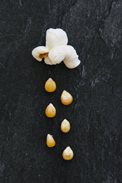 Konzept mit Popcorn und Mais - Popcorn regnet aus einem Popcorn - Wolke mit Regen als Popcorn und Mais vor schwarzem Hintergrund und rundherum Zuckerkörner - Foto, Bild