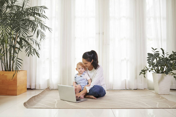 junge Frau sitzt mit Kind am Laptop auf dem Fußboden und beantwortet E-Mails auf dem Laptop - Foto, Bild