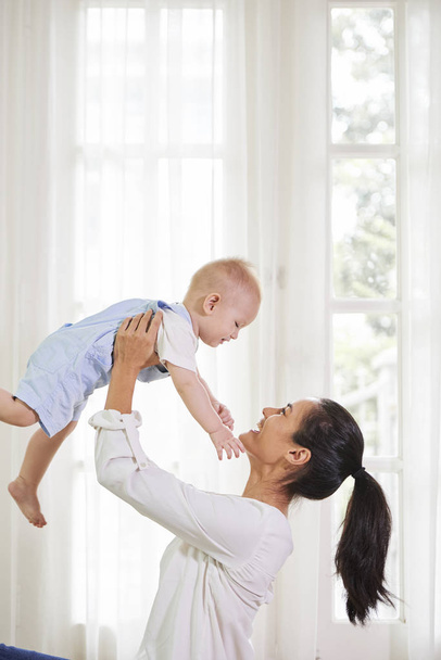 Heureuse jeune mère excitée soulevant son adorable bébé garçon souriant en coton dans l'ensemble lorsqu'elle se tient contre une grande fenêtre
 - Photo, image