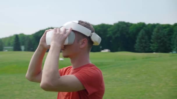 Człowiek stawia Gogle VR na głowie w parku i rozgląda się. Młody facet w czerwonym T-shirt i kask przeżywa wirtualną rzeczywistość na tle natury. Dynamiczne fotografowanie. materiał filmowy 4K. - Materiał filmowy, wideo