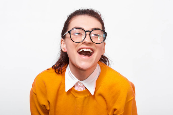 幸せな若い女性のクローズアップスタジオの肖像画は、広く微笑んで、オレンジ色のセーターと丸い透明の眼鏡を身に着けています。白いスタジオの壁の上にポーズのかわいい女子学生。人と感情 - 写真・画像