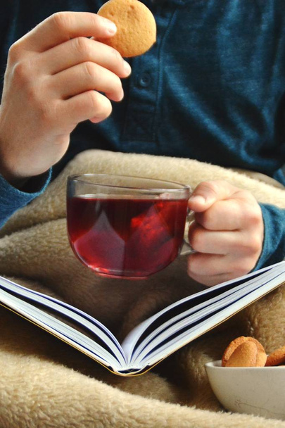 Un joven está sentado en un sofá y leyendo un libro mientras sostiene un té de frutas en una taza de vidrio en la mano y galletas a su lado - primer plano del libro y taza de vidrio llena de té - concepto para calentar
 - Foto, Imagen