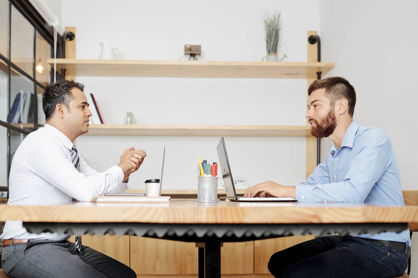 Dwóch biznesmenów siedzących przy stole z komputerami laptopami naprzeciw siebie i omawiając chwile pracy razem w biurze - Zdjęcie, obraz