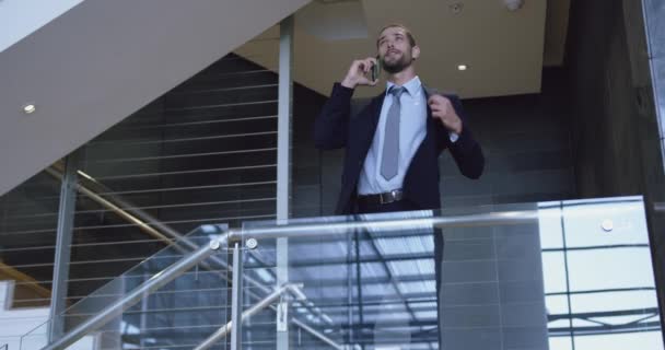 Front view of Caucasian businessman talking on mobile phone near staircase in office near railing. - Imágenes, Vídeo