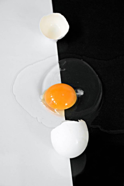 An opened egg with intact egg yolk surrounded by egg whites lies on a half black half white background - concept with black and white and an egg  - Foto, afbeelding