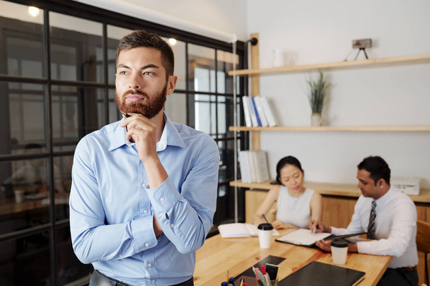 Young bearded businessman standing at office and thinking over something with his partners sitting at the table and working together in the background - Photo, image