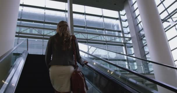 Low angle view of businesswoman moving upstairs on escalator in office and looking away. - Imágenes, Vídeo