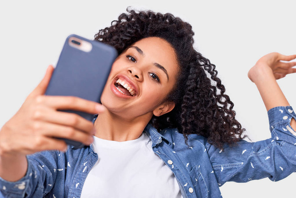 Closeup studio portrait of beautiful African American young woman in blue shirt, taking selfie over white studio wall. Happy African American female smiling, taking a self portrait on her smartphone. - Fotó, kép