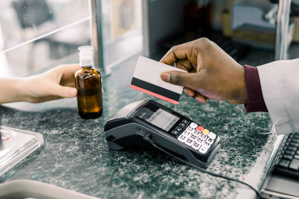 Cropped image of hands of African Man pharmacist holding credit card of client and hand of woman paying for Medicaments with credit card in pharmacy drugstore. - Фото, изображение