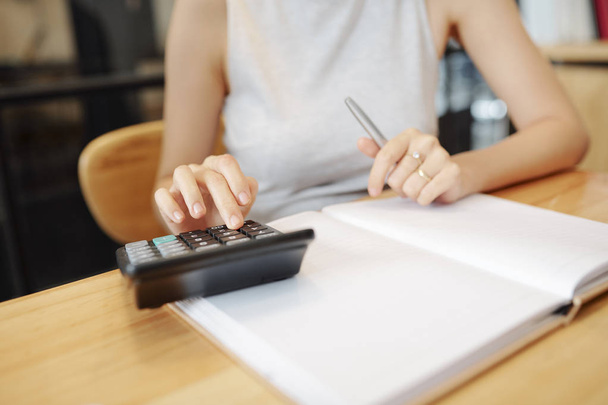 Close-up of businesswoman sitting at the table counting on calculator and taking notes in notebook - Foto, Imagem