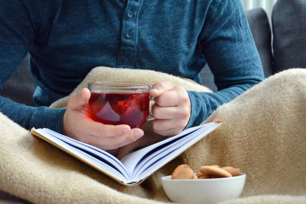 Un joven está sentado en un sofá y leyendo un libro mientras sostiene un té de frutas en una taza de vidrio en la mano y galletas a su lado - primer plano del libro y taza de vidrio llena de té - concepto para calentar
 - Foto, imagen