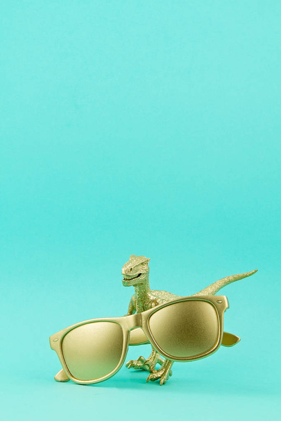 Brinquedo de dinossauro dourado segurando óculos de sol dourados. Tendências de verão, moda, vendas, ideia de compras
 - Foto, Imagem