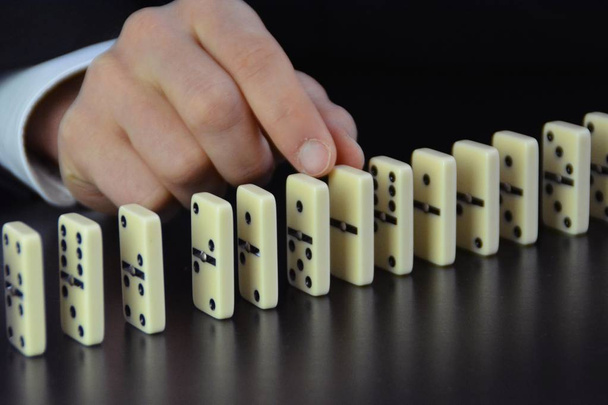 Close-up de uma mão de um jovem empresário examinando um fator de perturbação em uma reação em cadeia, de outra forma perfeitamente funcional, de dominós simbólico da complexidade das decisões de liderança
  - Foto, Imagem
