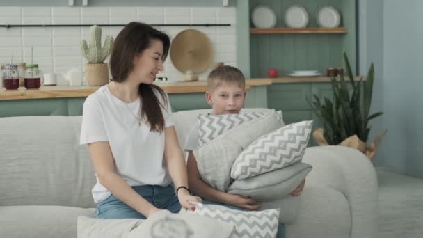 Family in shirts and jeans throw up pillows. Little son having funny pillow fight with mom on couch in living room. Son and mom playing together on bed at home in slow motion. - Felvétel, videó