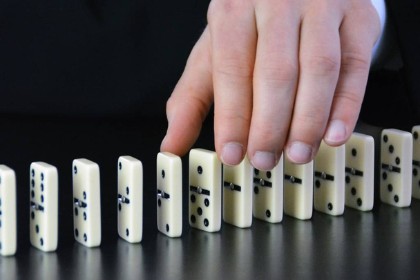 Close-up van een jonge zakenman die een verstoring factor onderzoekt in een anderszins perfect functionerende kettingreactie van domino's-symbolisch voor de complexiteit van leiderschaps beslissingen  - Foto, afbeelding