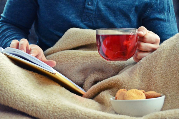 A young man is sitting on a sofa and reading a book while holding a fruit tea in a glass cup in hand and biscuits stand beside him - closeup on the book and glass cup full of tea - concept to warm up - Photo, Image