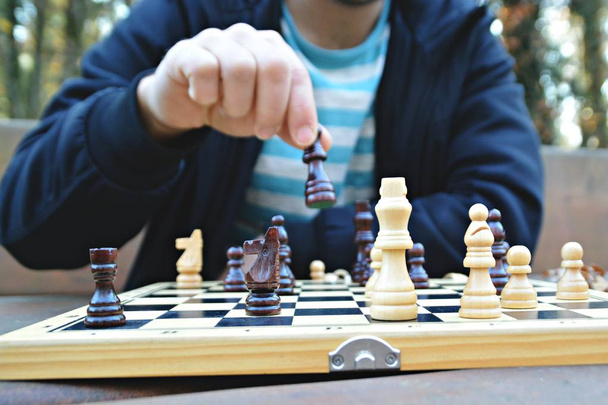 Um homem de vinte e poucos anos senta-se em um parque outonal e joga xadrez focando nas peças de jogo no tabuleiro, a pessoa não pode ser vista e apenas as mãos e o tronco são visíveis
 - Foto, Imagem