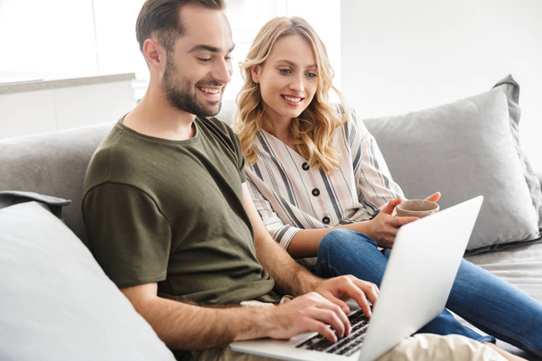 Happy excited young loving couple sitting on sofa at home using laptop computer. - 写真・画像