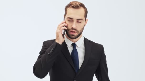 Serious young businessman in black suit and blue tie becoming angry and gesturing with hand while having a call over gray background isolated - Filmagem, Vídeo