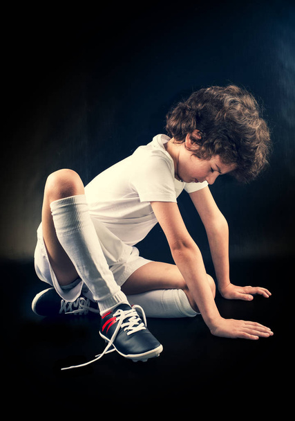 Little disappointed boy soccer player sitting on the floor with untied lace on the boots. Dark background - Фото, зображення