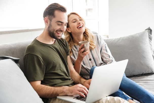 Happy excited young loving couple sitting on sofa at home using laptop computer. - 写真・画像