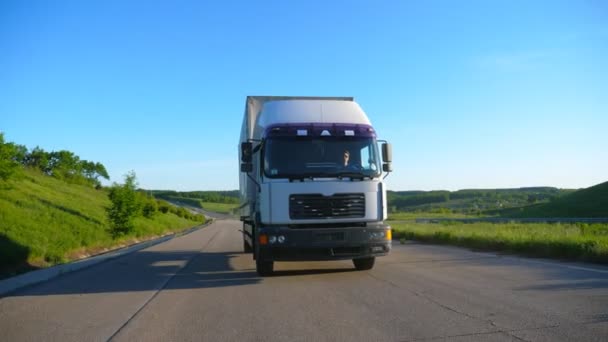 Vooraanzicht van vrachtwagen met vrachtwagen met snelheidsoverschrijding op de snelweg en goederenvervoer op zonnige dag. Witte vrachtwagen rijden door landelijke weg met prachtige natuur landschap op de achtergrond. Slow Motion - Video
