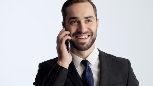 Happy young businessman in black suit and blue tie smiling and laughing while having a call on smartphone over gray background isolated - Materiał filmowy, wideo