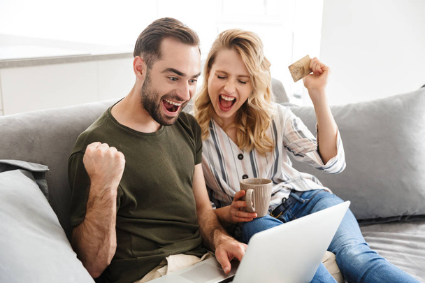 Happy excited young loving couple indoors at home sitting on sofa using laptop computer make winner gesture holding credit card. - Photo, image