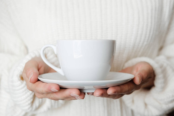 Woman in white knitted sweater holding cup of coffee. Female hands with cup of tea. Close up - Фото, зображення