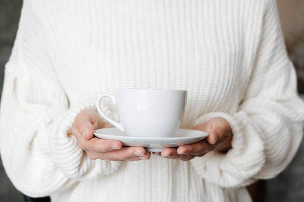 Жінка в білому в'язаному светрі тримає чашку кави. Жіночі руки з чашкою чаю. крупним планом
 - Фото, зображення