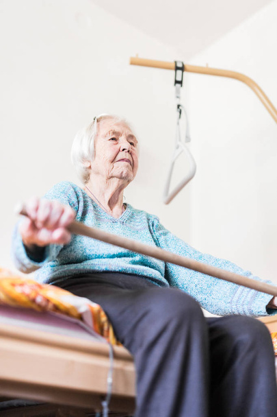 Elderly 96 years old woman exercising with a stick sitting on her bad. - Foto, Bild