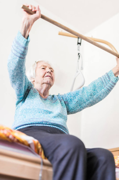 Elderly 96 years old woman exercising with a stick sitting on her bad. - Φωτογραφία, εικόνα