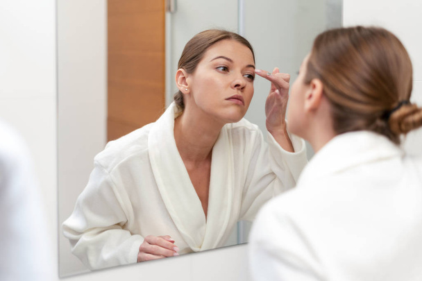 Woman in bathrobe looking in mirror at bathroom - Zdjęcie, obraz