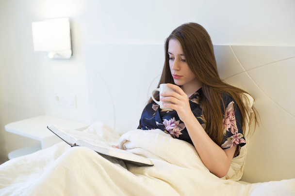 young attractive brunette woman lying in bed wearing silk dressing gown holding cup of offee and reading book - Photo, Image