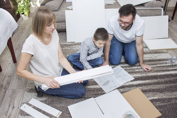Dad and son are staring at the furniture assembly instructions kneeling on the carpet. Mom chooses one element from many that lie on the carpet. - Фото, зображення