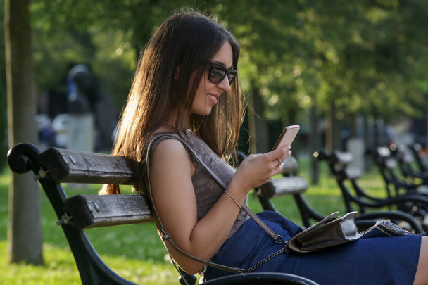 Frau tippt Nachricht auf Smartphone, sitzt auf Parkbank - Foto, Bild