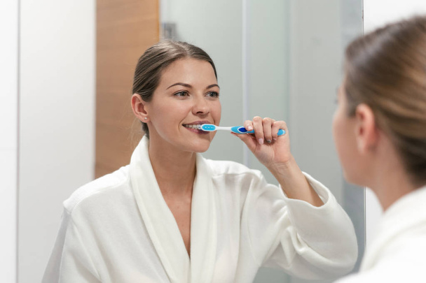 Femme en peignoir blanc tenant une brosse à dents, debout dans la salle de bain
 - Photo, image
