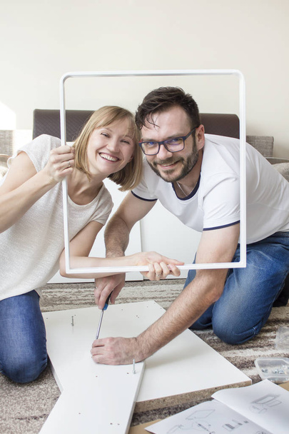 Mann und Frau verdrehen Möbel im Wohnzimmer. Lächelnde Menschen posieren für ein Foto mit einem Tischelement als Fotorahmen. - Foto, Bild